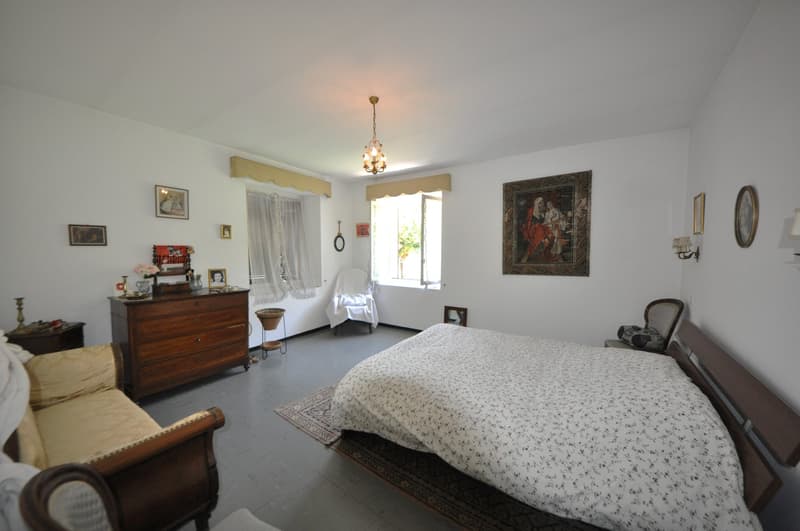 Bedroom with a bed, couch, wooden dresser, chandelier, picture frames, lamps, small table, and window with curtains.