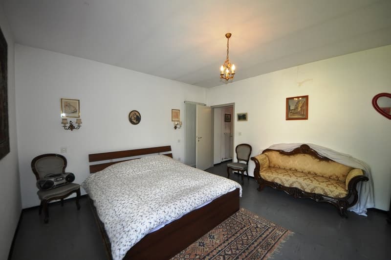 Spacious bedroom with a wooden bed frame, a vintage-style sofa, and a chandelier overhead. The room features white walls, hardwood floors, and various decorative elements such as framed artwork and wall sconces.