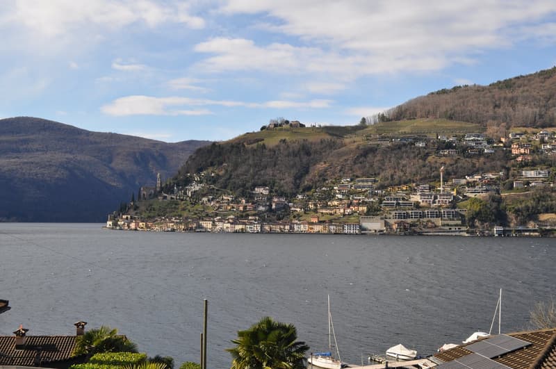 Scenic view of a lakeside town nestled in the hills, with buildings and houses scattered along the slopes. The town is situated on the shore of a large body of water, likely a lake or river. The landscape is dominated by forested hills and mountains in th