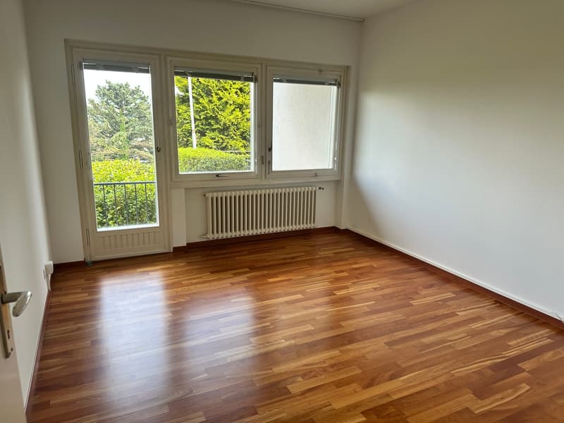 Spacious bedroom with hardwood flooring, large windows overlooking a lush green outdoor area, and a radiator heating system.