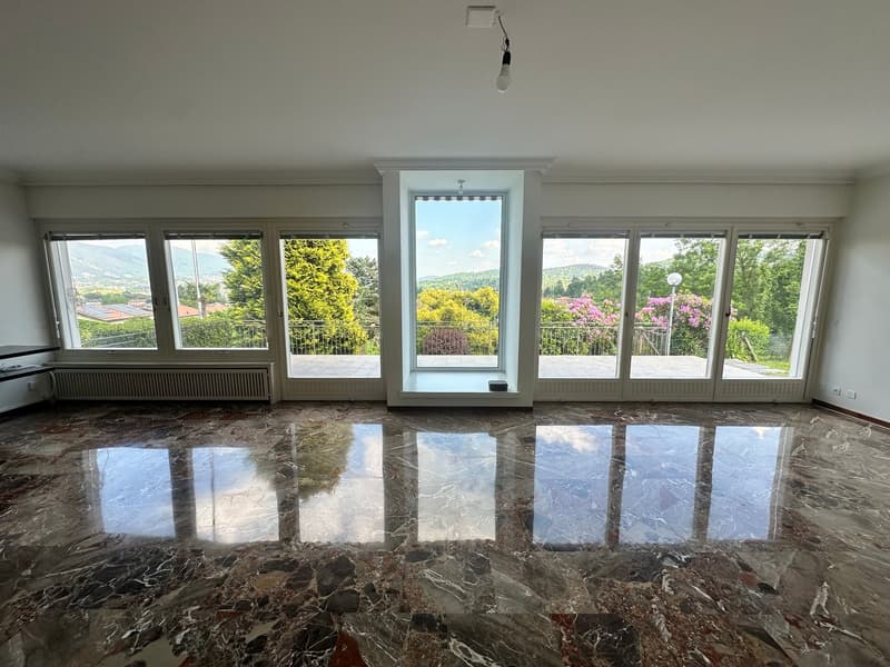 Spacious living room with large windows offering a scenic view of the surrounding nature, including trees and mountains. The room has a marble floor with a polished, reflective surface.