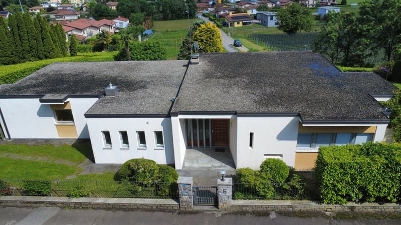 Two-story modern house with a flat roof, white exterior walls, and yellow accents. The house has a garage and is surrounded by a well-maintained garden with trees and shrubs. The property is situated on a hill overlooking a residential neighborhood.