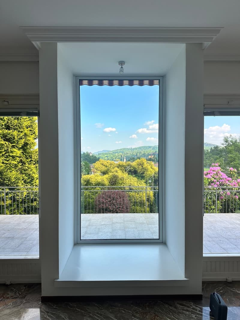 Large window with a scenic view of the surrounding greenery and mountains, tiled floor, white walls, and a decorative molding around the window frame.