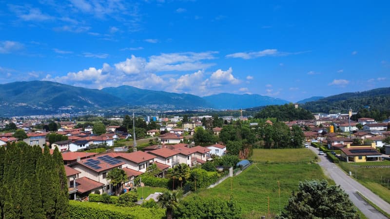 The image shows a residential area with a mix of single-family homes and multi-unit buildings. The houses have red tile roofs and are surrounded by lush greenery, including trees and gardens. The area is situated in a valley, with mountains visible in the