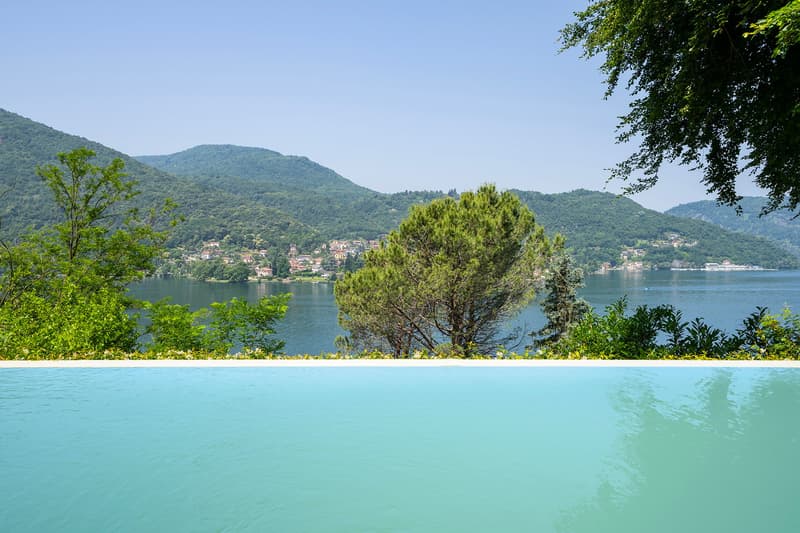 The image shows a scenic view of a lake surrounded by lush green mountains and forests. The lake has a turquoise blue color and is framed by a sandy beach and pine trees in the foreground. The landscape appears to be a tranquil and picturesque setting.