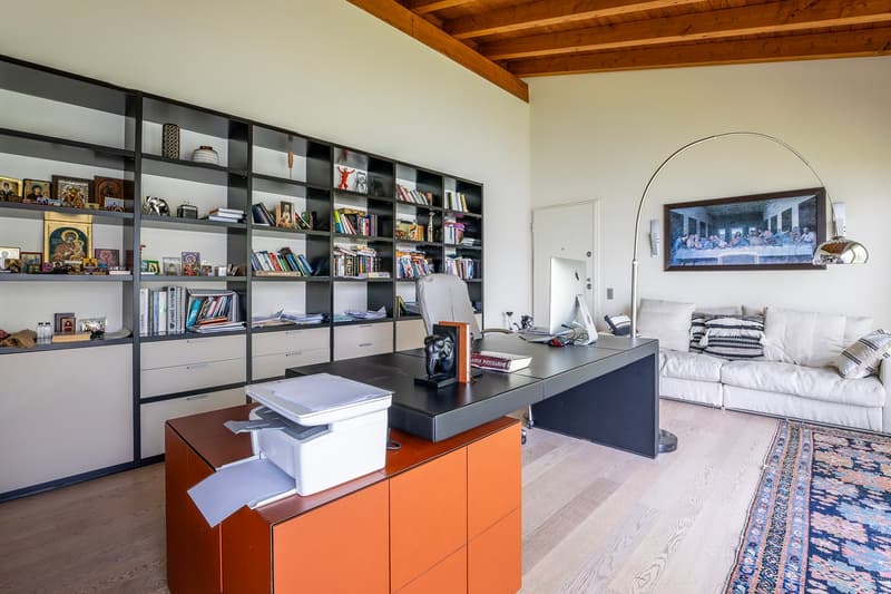Spacious home office with built-in bookshelves, a large desk, and a couch in the living area. The room has wooden beams on the ceiling and a large TV on the wall. There are various decorative items and books on the shelves.