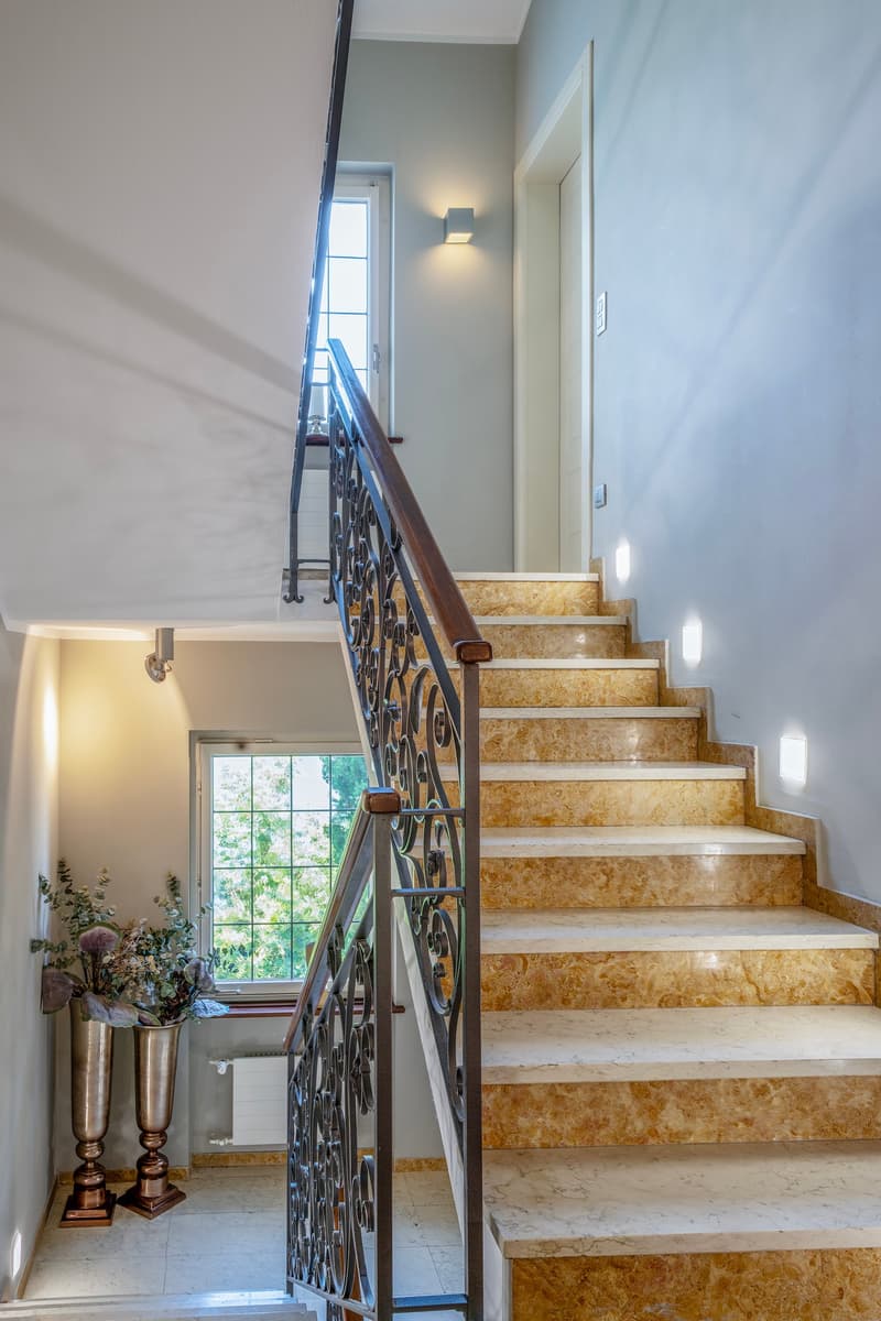 Marble stairs with ornate metal railing, wall sconce lighting, potted plants at the bottom of the stairs