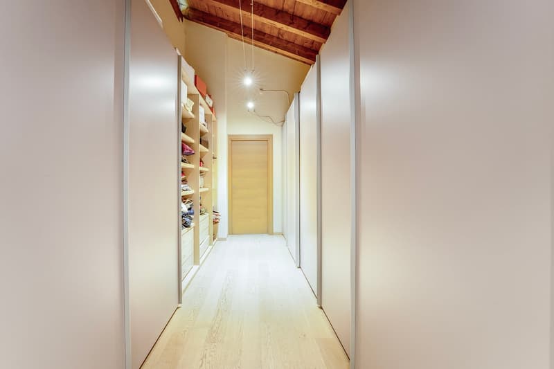 Long hallway with white walls, wooden ceiling beams, and built-in shelving units on one side. The floor is made of light-colored wood planks. The hallway leads to a wooden door at the end.