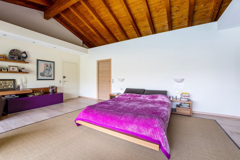 Spacious bedroom with wooden beamed ceiling, white walls, hardwood floor, large bed with purple bedding, wooden nightstands, and a purple cabinet/entertainment unit.