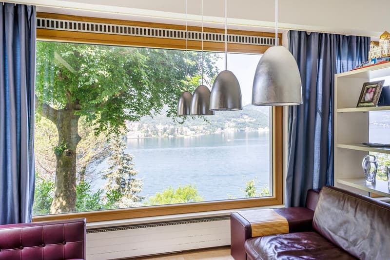 Large window with a scenic view of a lake surrounded by trees, leather sofa, pendant lighting fixtures, wooden window frame