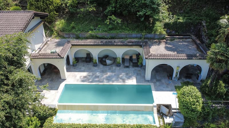 Two-story Mediterranean-style villa with arched windows and balconies, surrounded by lush greenery. The property features a large swimming pool and a covered outdoor living area with seating.