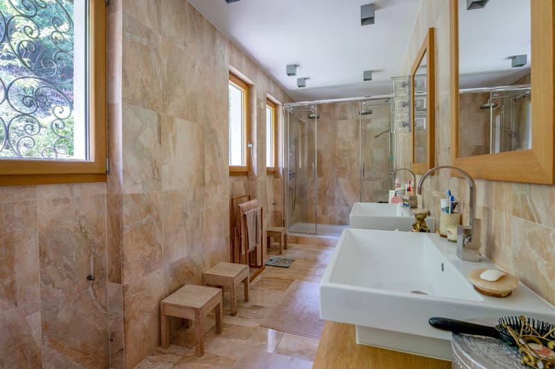 Large bathroom with stone tile walls, wooden accents, glass shower enclosure, vanity with sink, and a window overlooking a balcony or terrace area.