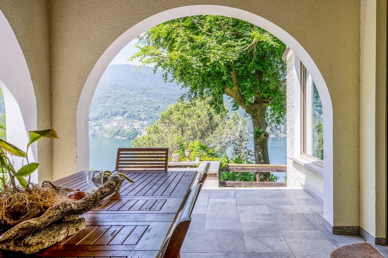 Arched doorway leading to a wooden deck with a table and chairs, overlooking a scenic lake and mountains in the distance. A large tree provides shade and greenery to the terrace.