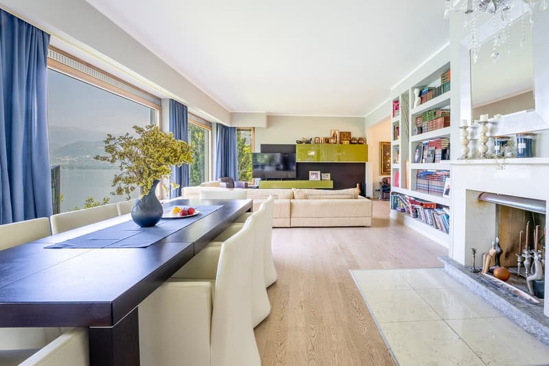 Spacious living room with large windows offering a scenic view, hardwood flooring, a long dining table, a comfortable sofa set, a TV unit with shelves, and a chandelier overhead.
