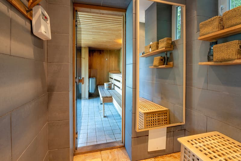 The image shows a laundry room with a tiled floor, wooden shelves, and a washing machine. There are woven baskets and other storage containers on the shelves. The room has a simple, rustic design with a wooden door frame and exposed wooden beams on the ce