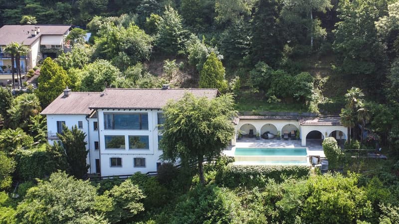Two-story house with a blue exterior, large windows, and a swimming pool surrounded by lush greenery and palm trees. The house also has a covered terrace or balcony area.