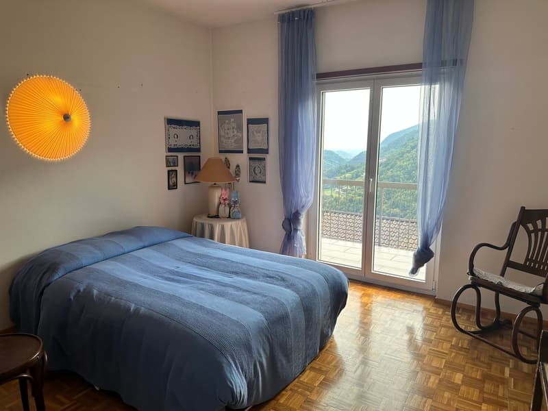 bedroom with bed, side table with lamp, rocking chair, window with curtains, balcony with railing, wall decorations