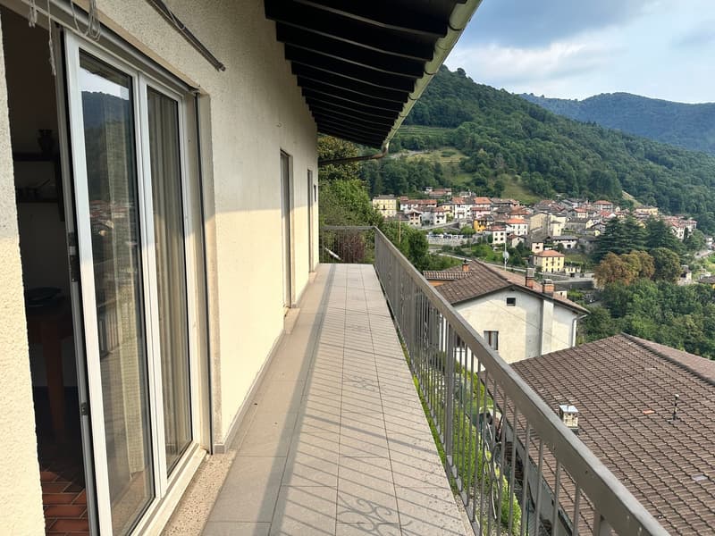 Balcony with a metal railing and tiled floor, overlooking a mountainous village scene with greenery and houses.