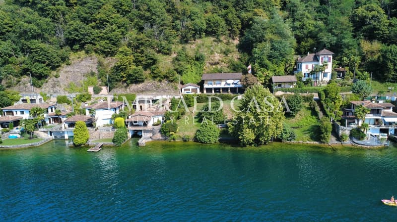 Multiple houses on a hillside overlooking a lake, stonework and brick exterior, green water, surrounded by trees and plants, a small boat in the foreground