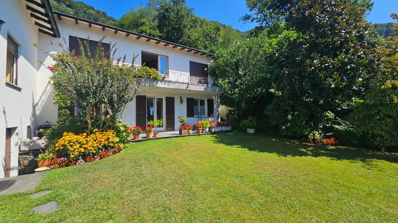 2 story house, white exterior, wooden doors, balcony with railing, flower-filled yard, well-manicured lawn, multiple flower pots