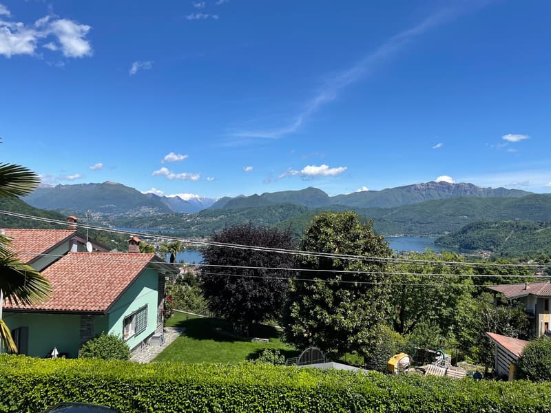 House with tiled roof, green house in the foreground, greenery, trees, clear blue sky, mountains in the distance, lake, elevated view