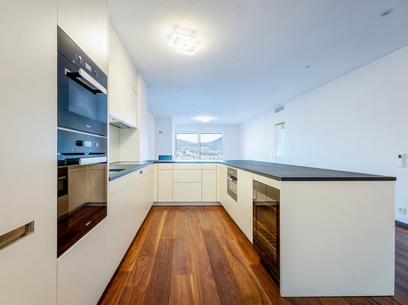 Modern kitchen, white cabinets, granite countertops, integrated oven and dishwasher, wooden floor, ceiling lights, and large window with mountain view.
