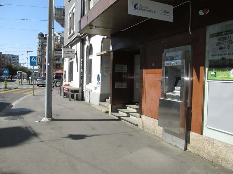 Building exterior with ATM, steps leading to entrance, sign for Zurich Kantonalbank, benches, and a streetlight.