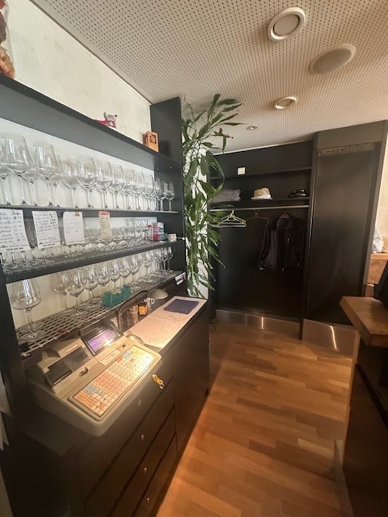 A room with a wooden floor, a black shelf with wine glasses, a plant, a desk with a computer, a cash register, and a wooden table.