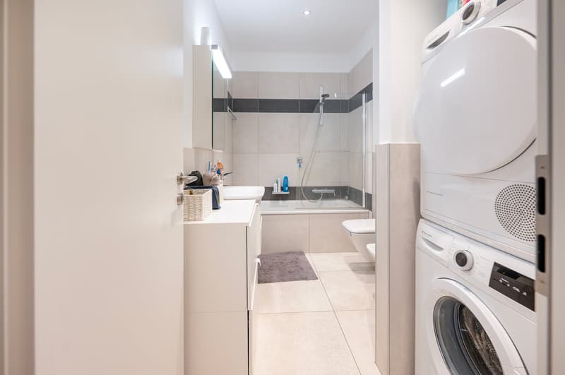 a modern white bathroom with white tiles, a large bathtub, a toilet, a vanity, a basket with towels, a shower, and a washing machine