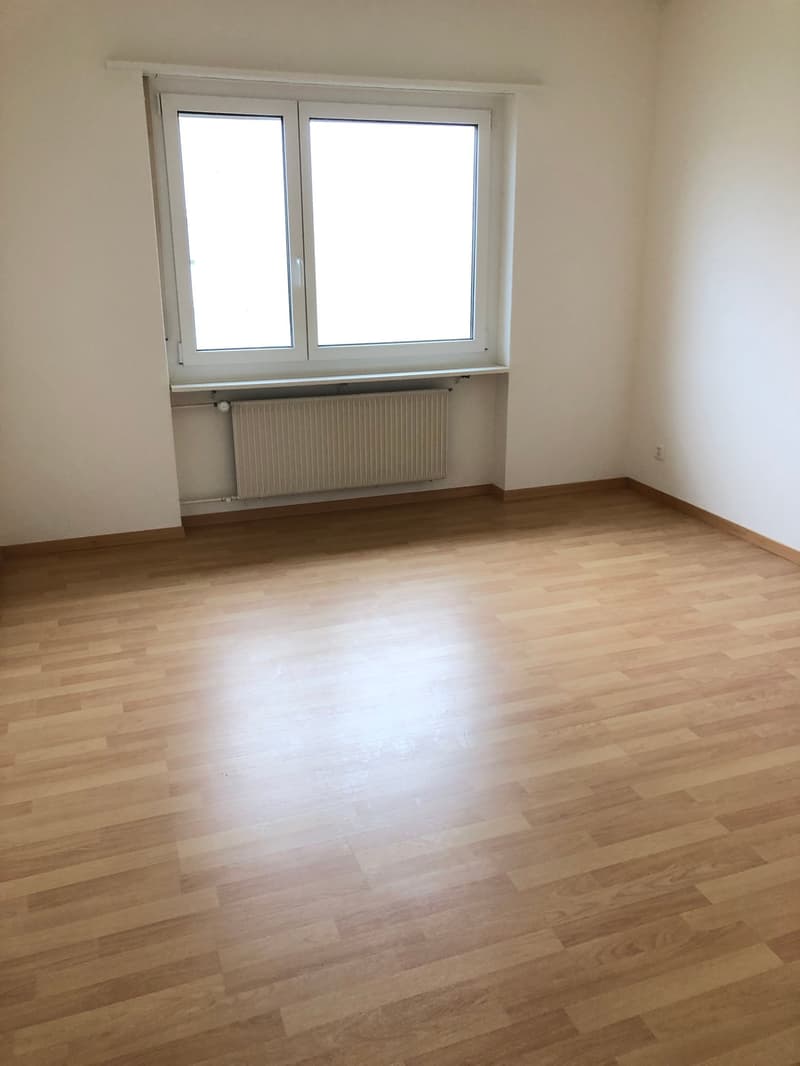 The image shows a spacious bedroom with large windows, hardwood flooring, and a radiator on the wall.