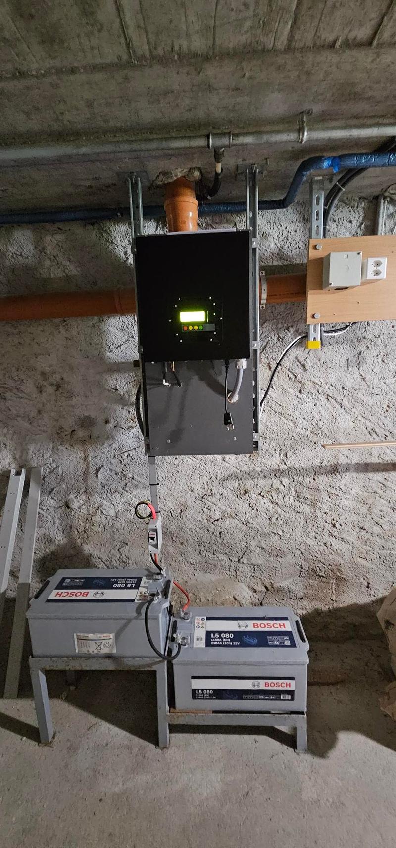 Two Bosch batteries, connected to a black central unit with a yellow and green indicator light. Metal stands, wires, pipes.