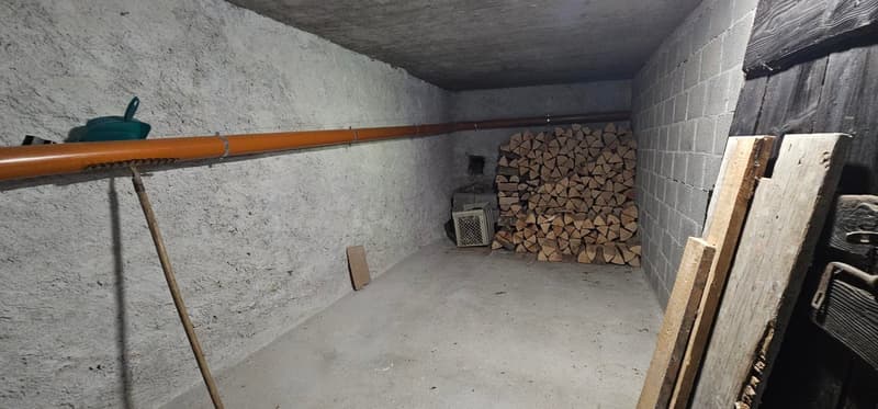 A concrete room with a pile of wood against a brick wall, a small furnace-like machine, and a large wooden stick.
