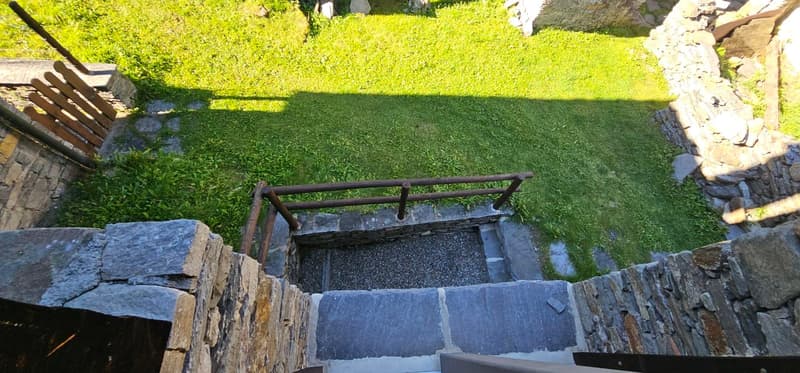 Staircase with stone walls, leading to a raised ground floor and surrounded by a grassy lawn.