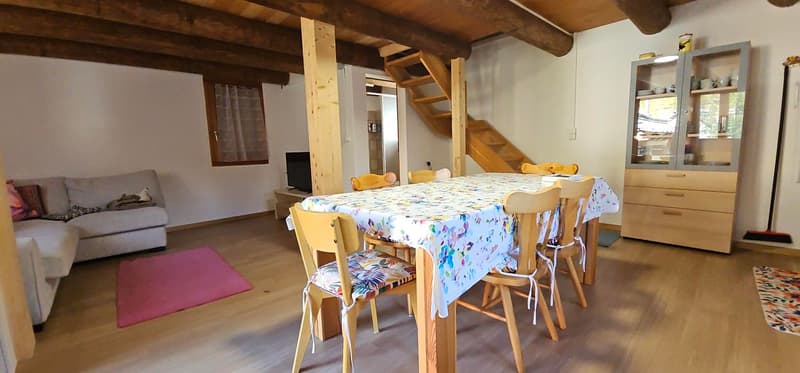 Living room, dining area with wooden dining table and chairs, couch with cushions, stairs to the upper floor, shelf with glass doors