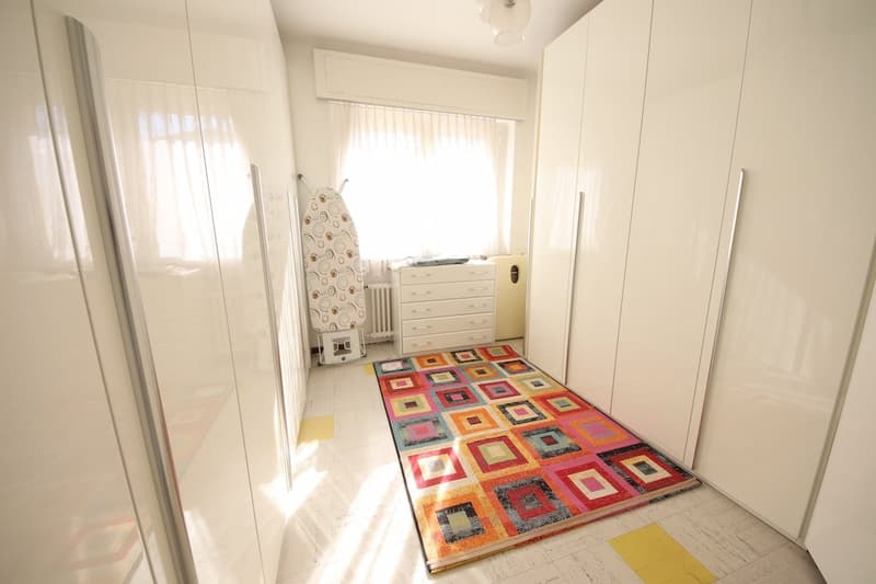 white walls, tiled floor, colorful rug, wardrobes, large window, ironing board, cabinet