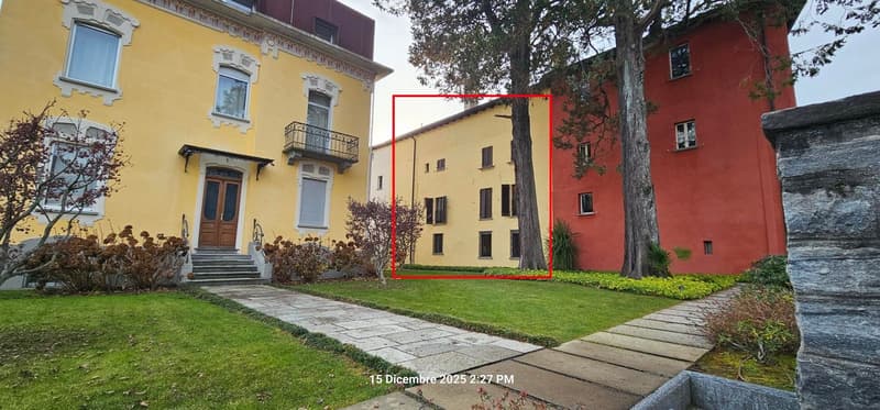 Facade of a yellow building with red frame, steps to the door, balcony, garden and trees in front.