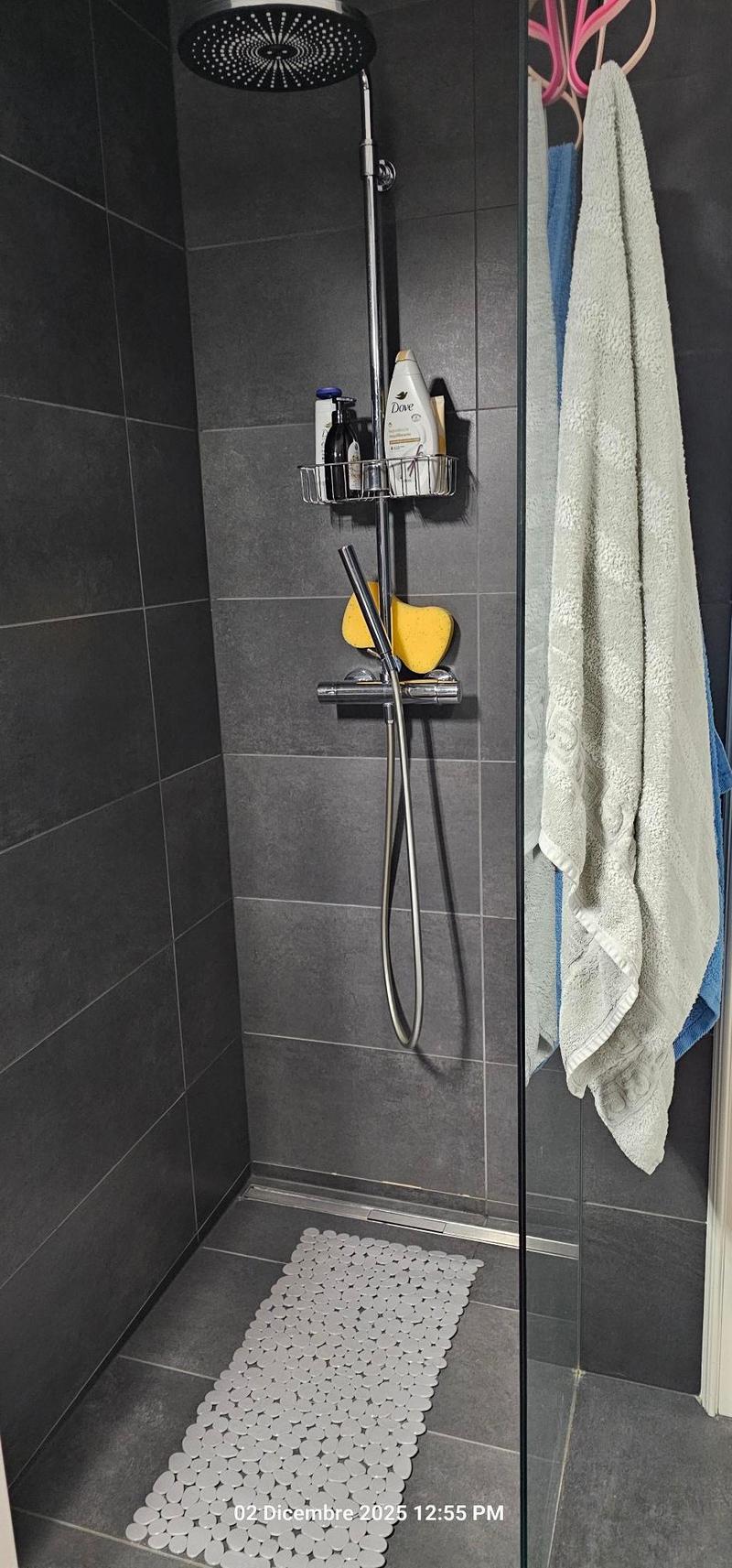 walk-in shower with silver showerhead, towel hanging on rack, glass door, tiled floor, gray tiled walls, floor mat, product shelves