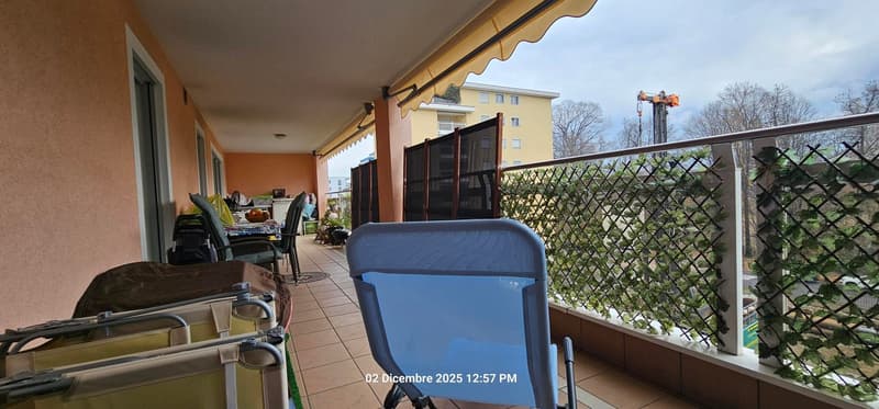 Terrace with awning, chairs, potted plants, metal railings, tiled floor, distant buildings