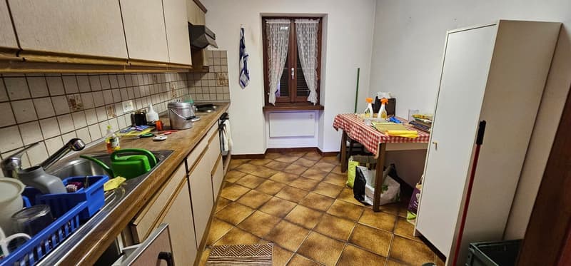 tiled floor, tiled walls, kitchen counter, sink, sink faucet, gas stove, wooden cabinets, electrical outlets, window, door, white cupboard, table, bags