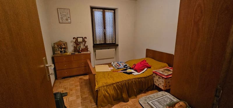 Empty bedroom with wooden bed, wardrobe, bed side table, small table on right, wooden cabinet with drawers on left, framed picture above cabinet