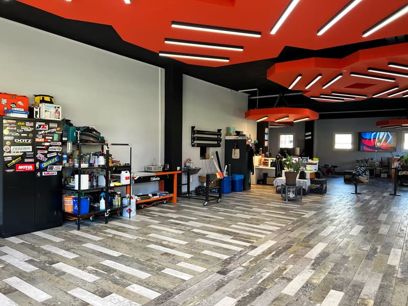 Workshop with orange ceiling, large wooden table, shelving unit, cleaning products, tools, storage boxes, monitors, blue bins, potted plant, empty desk