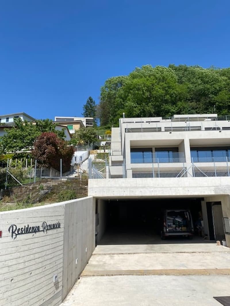 Multi-story apartment building with a garage and parking spaces on the ground level, surrounded by trees and greenery.