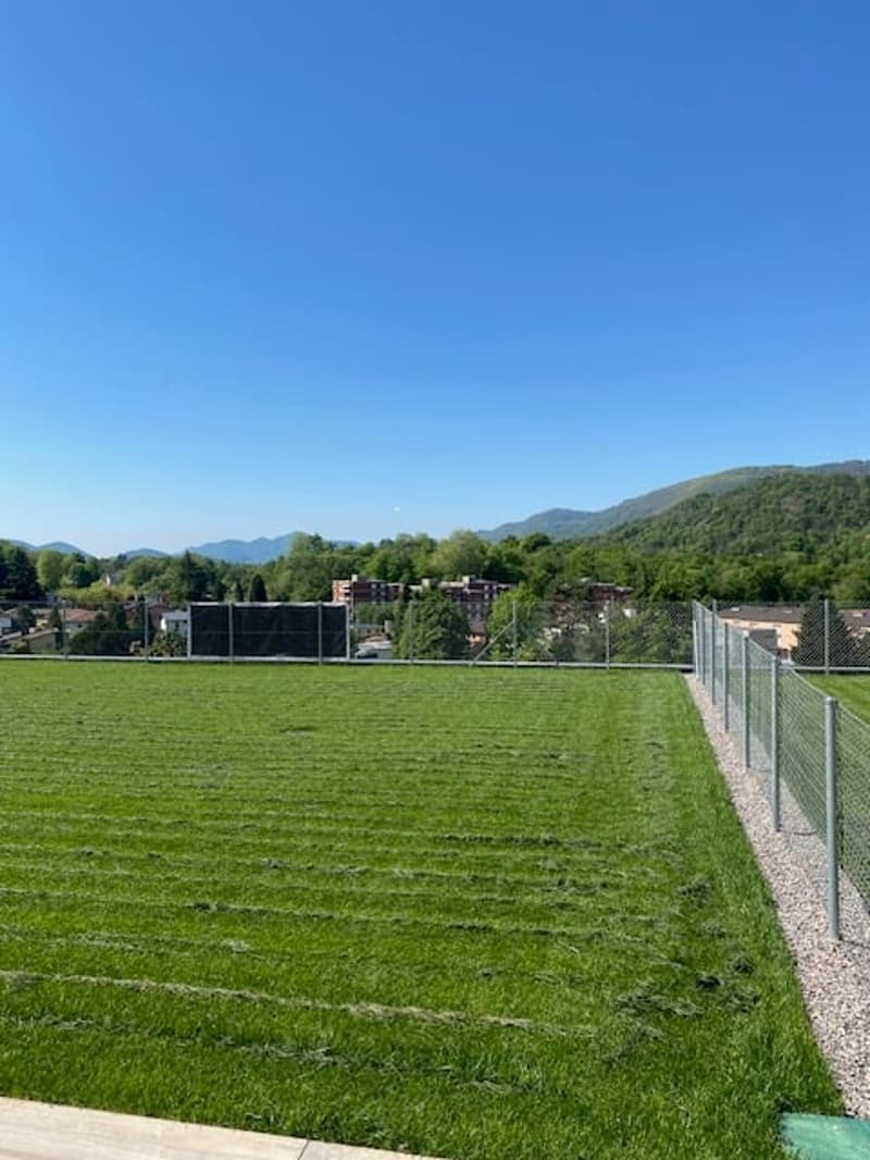 The image shows a large grassy field surrounded by a wire fence. The field appears to be a sports field or playing area, with a clear, well-maintained grass surface. In the background, there are mountains and a blue sky, indicating a scenic outdoor settin
