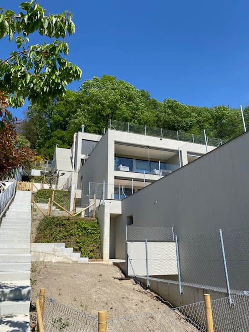 Modern multi-story house with a balcony, surrounded by lush greenery and trees