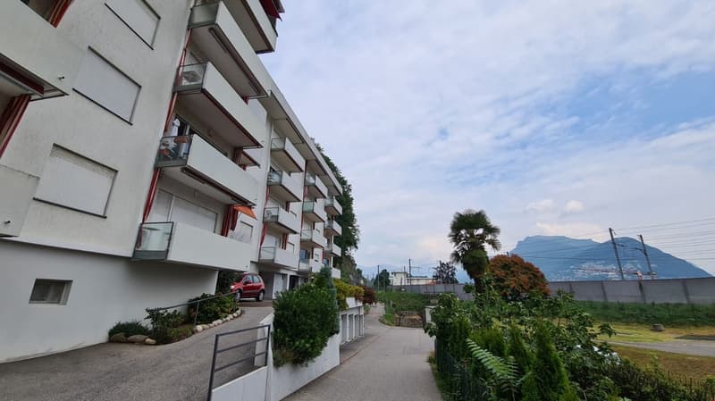 4-story building, modern design, balconies, white exterior, red car parked in front, mountain view in the background, clear sky, street lined with plants, pathway