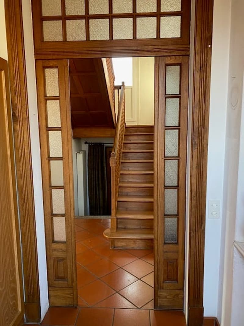 Wooden door, tiled floor, staircase, brown wooden banister, white wall