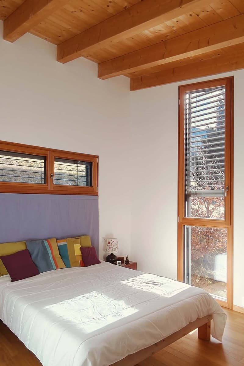 Bedroom with wooden bed, white bedding, wooden nightstand, lamp, and large windows with blinds