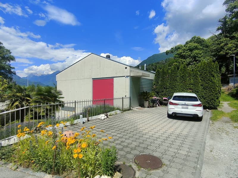 2-story building with a gray exterior, a red garage door, and surrounded by lush greenery and flowers. There is a paved driveway with a parked car in front of the building.