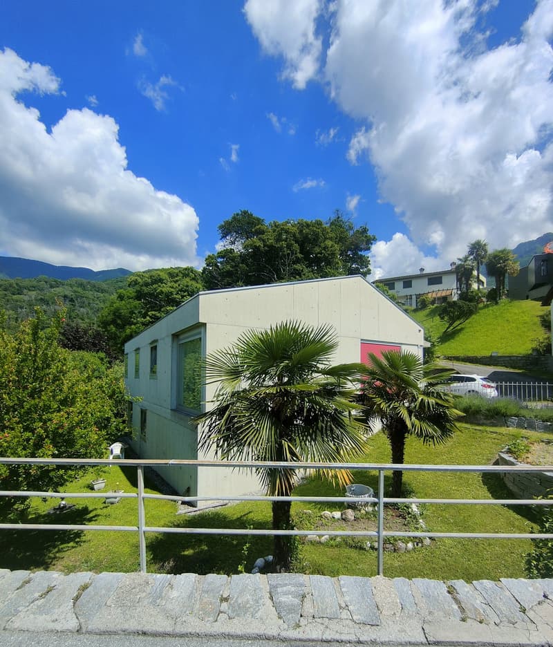 Single-story house with a flat roof, surrounded by lush greenery and palm trees. The house has large windows and a balcony or terrace overlooking the scenic landscape with mountains in the background.