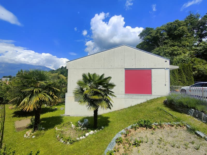 A modern, single-story building with a gray exterior and a large red door. The building is surrounded by lush greenery, including palm trees and other plants. There appears to be a parking area or garage adjacent to the building.